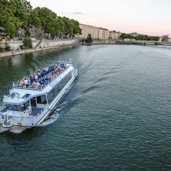 Turbåt med passagerare som kryssar längs en flod, med träd och byggnader som kantar flodbanken, och en bro i bakgrunden.