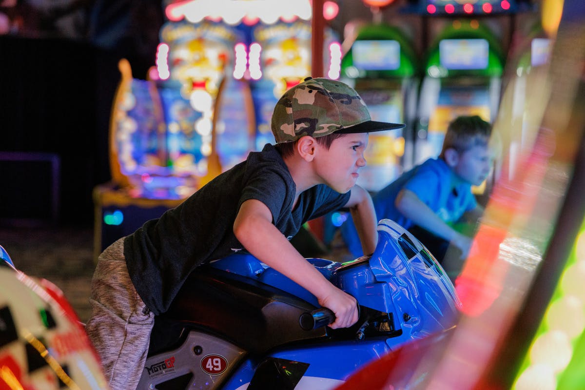 Two boys intensely focused while playing motorcycle racing arcade games in a brightly lit game arcade.