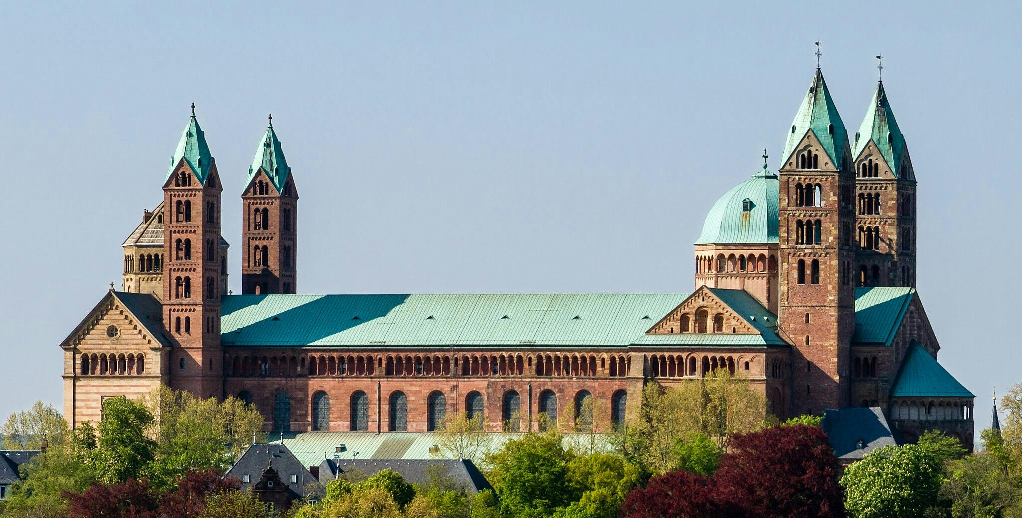Large medieval cathedral with multiple towers and green rooftops, surrounded by trees and buildings. Clear blue sky in the background.