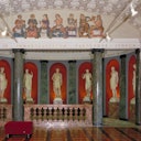 Ornate room with marble columns, red walls, and a series of classical statues. Ceiling fresco depicts various figures.