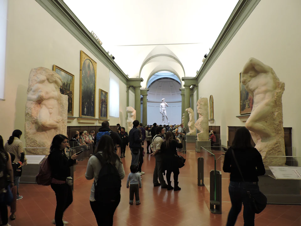 Visitors at a museum's sculpture hall, with large statues on both sides and a prominent white statue at the end of the hall.