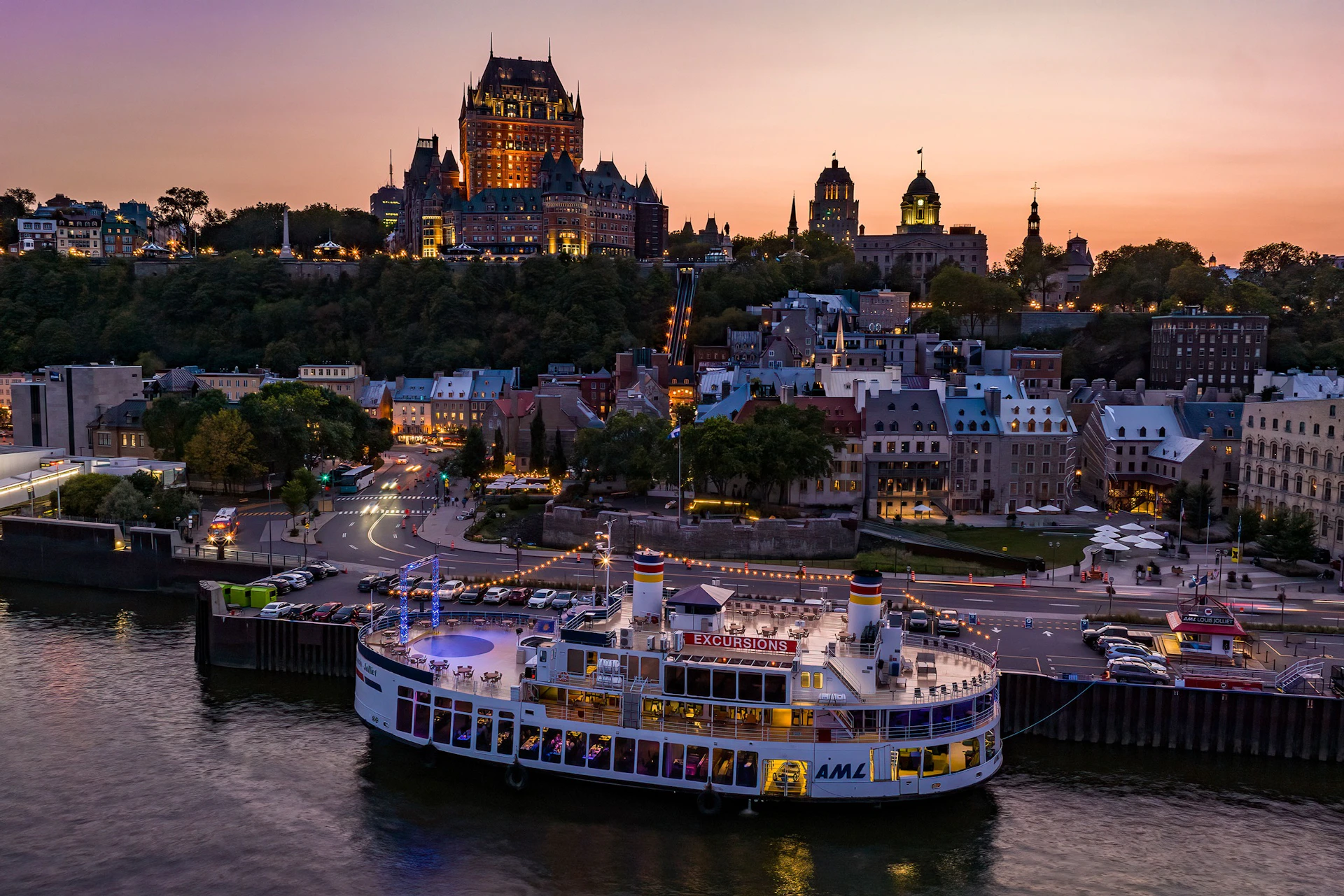 A brightly lit ferry on a river with a historic cityscape and illuminated buildings on a hill at dusk in the background.
