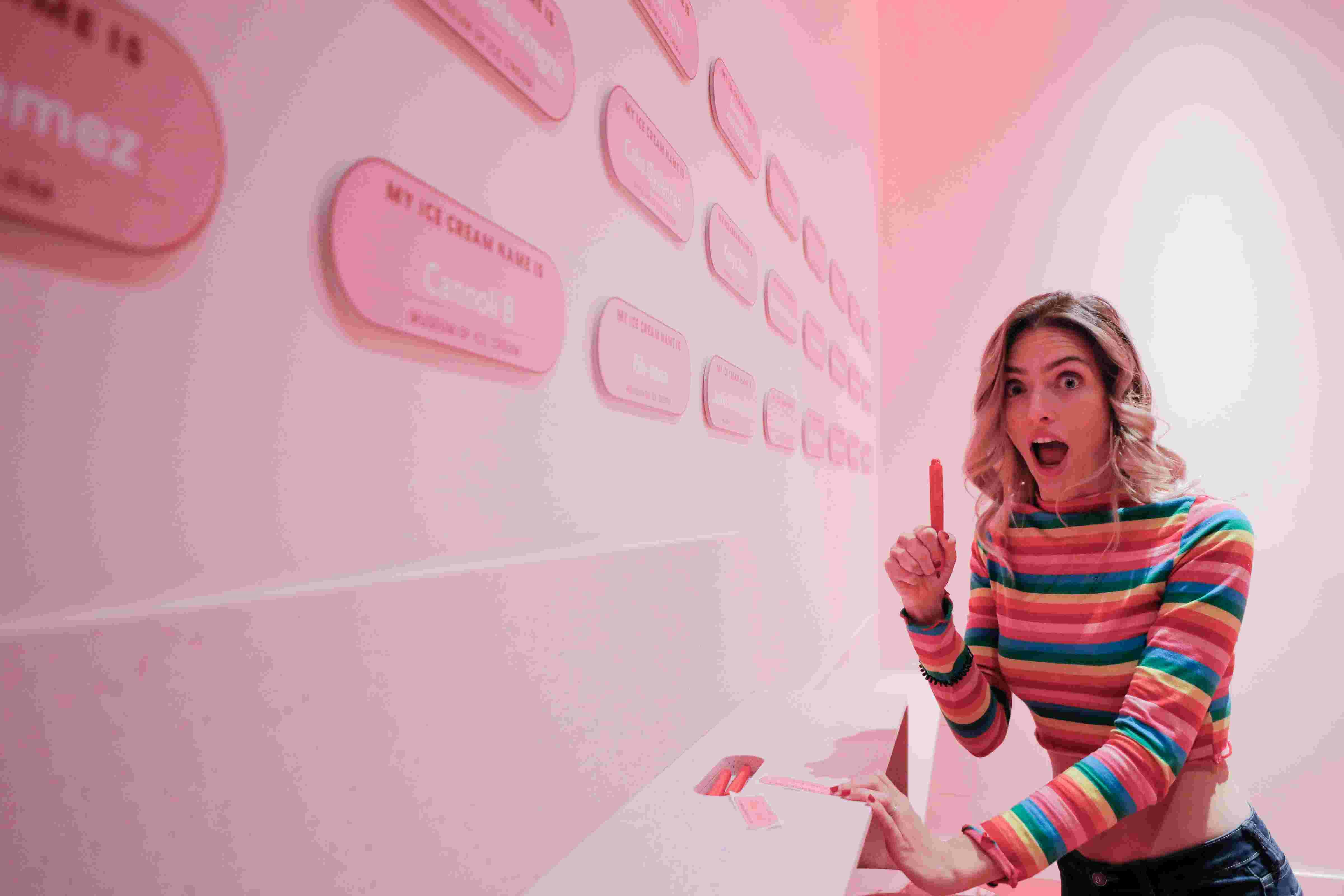 A surprised woman in a colorful striped shirt holds a popsicle while interacting with a wall featuring multiple labeled pink tags.