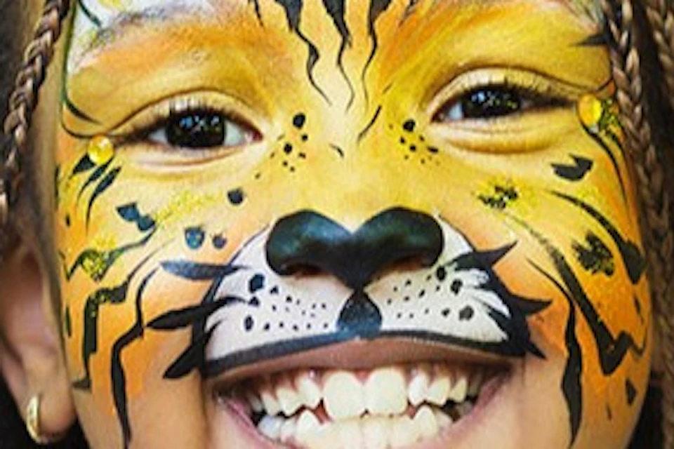 Child with face painted like a tiger, smiling and posing with hands raised, against a blurred green background.