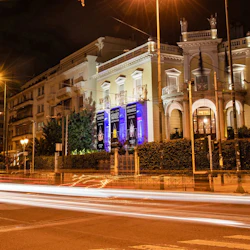 En ljust upplyst historisk byggnad på natten med ljusspår från förbipasserande bilar på gatan framför.