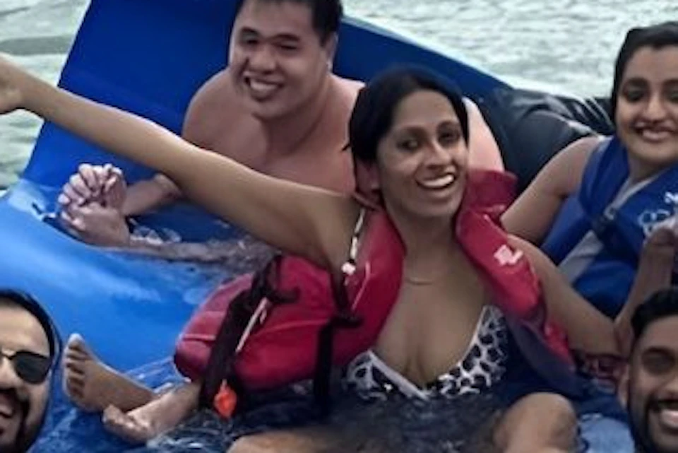 Six people relaxing on a blue floating mat in the water, holding drinks and making gestures, enjoying a sunny day.