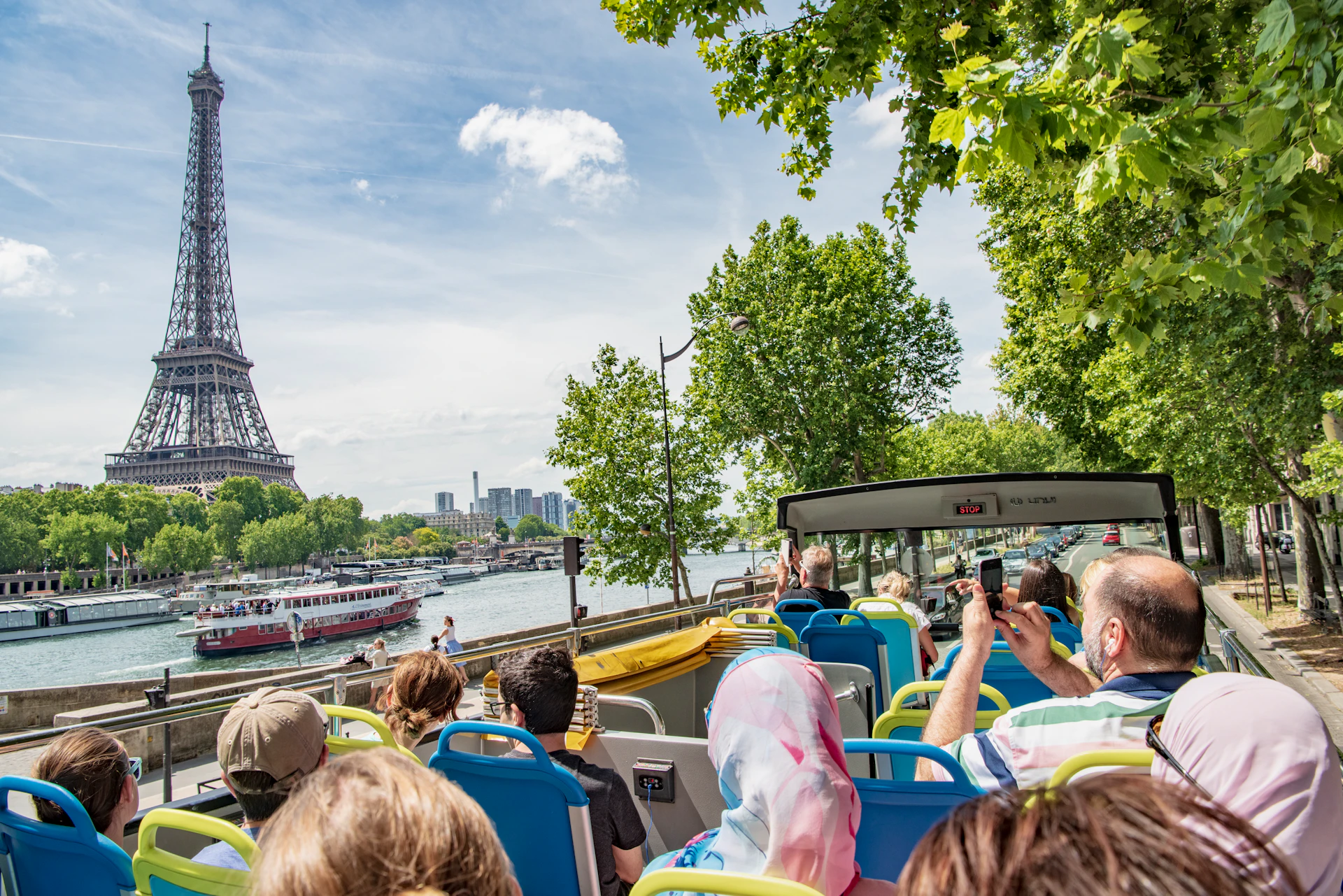 Tourists on an open-top bus tour near the Eiffel Tower and Seine River in Paris, with trees and other boats visible.