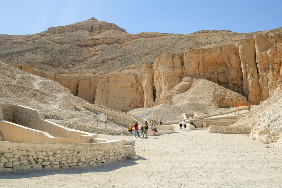 People walk among ancient limestone structures and pathways set against a desert mountain backdrop under a clear blue sky.
