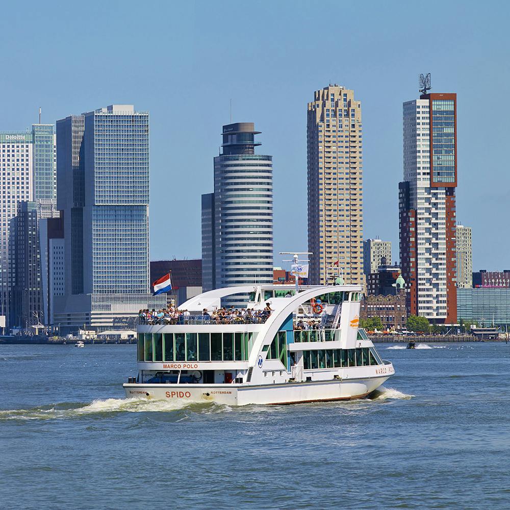 상부 갑판에 있는 사람들과 함께 강에서 즐기는 투어 보트. 맑고 푸른 하늘 아래 배경에 현대적인 고층 건물이 보입니다.