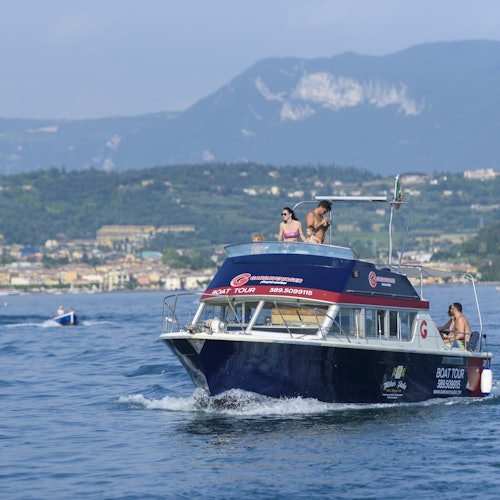 A boat with passengers on a lake, with mountains and a town visible in the background under a clear sky.