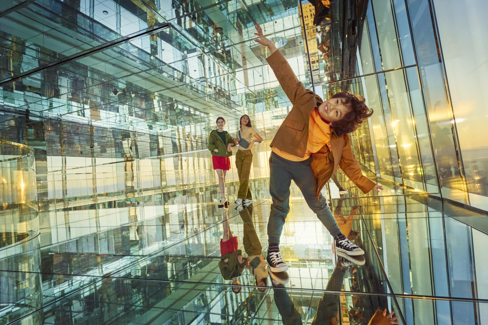 Tři lidé ve skleněné a zrcadlové místnosti, jedno dítě v popředí hravě pózuje.