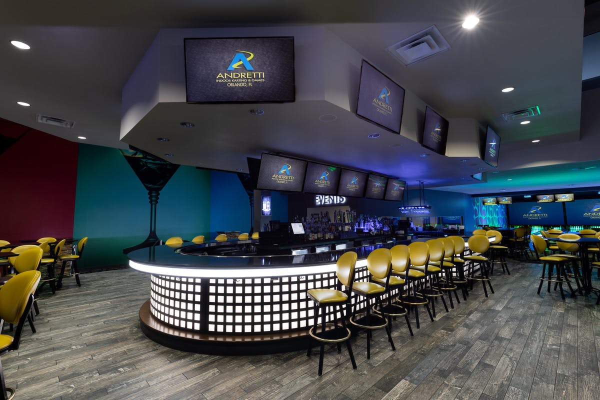 A modern bar with yellow stools, a curved counter, multiple screens displaying "Andretti," and a dim, ambient lighting.
