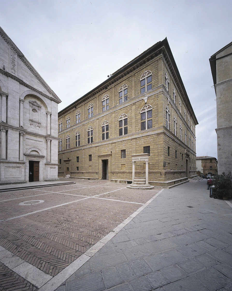 A cobblestone square with a large, ornate stone building on the left and a multi-story, arched window building on the right.