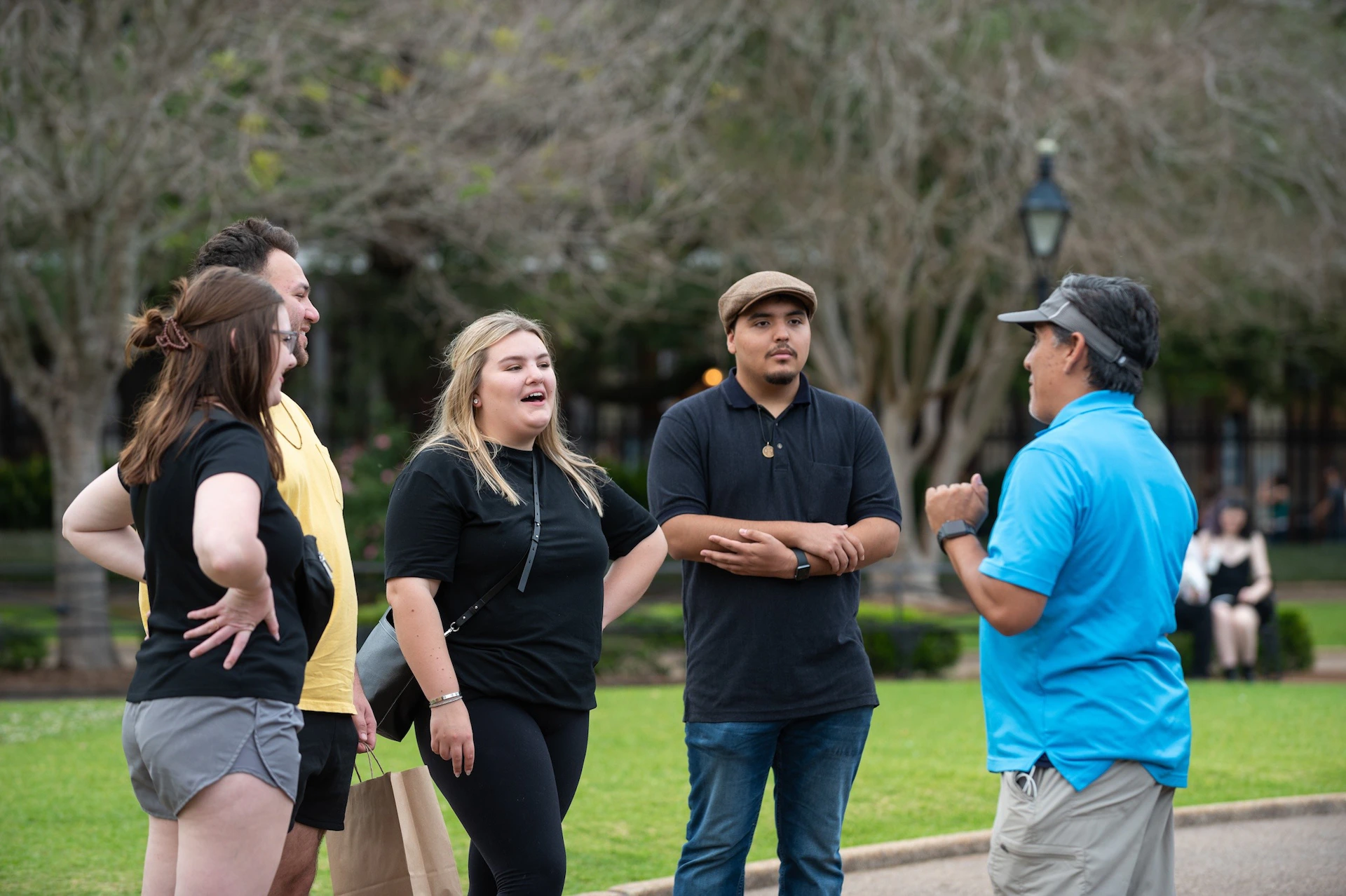 Three individuals are standing outdoors, engaged in a conversation with a man wearing a blue shirt and a cap.