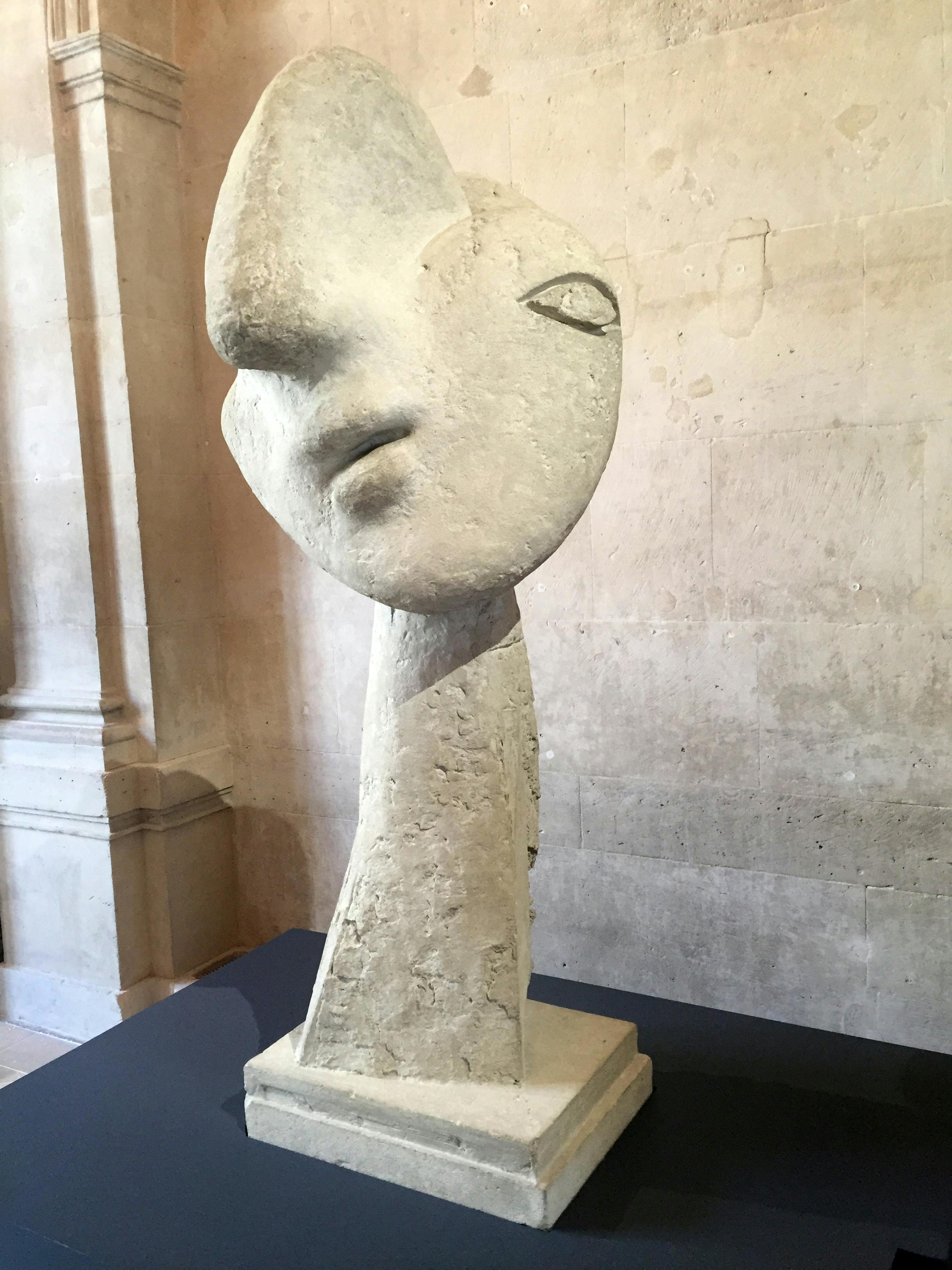 Abstract stone sculpture of a head with exaggerated features, displayed indoors against a beige wall with an architectural detail.