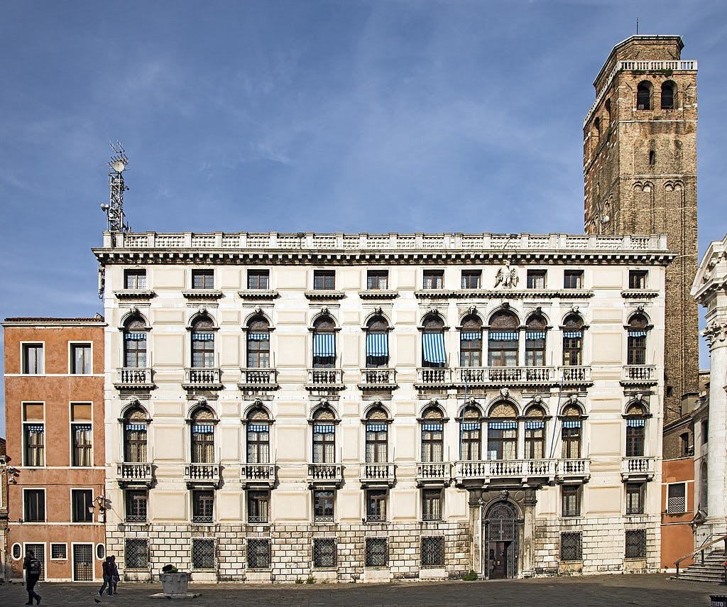 Elegant, multi-story historic building with arched windows, balconies, and an adjacent brick tower under a blue sky; several people walk by.