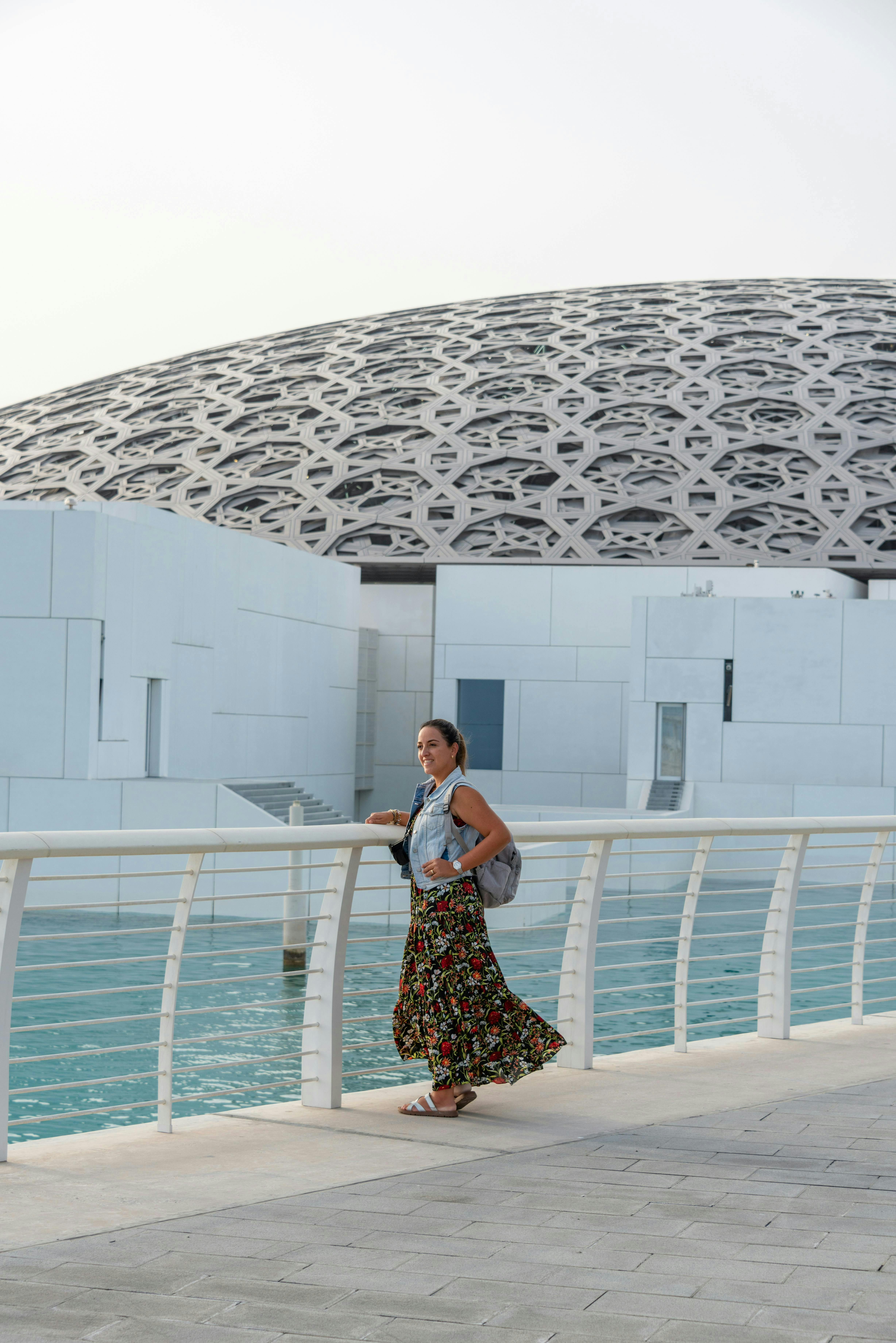 A woman in a floral skirt stands near a white railing, with water and a geometrically patterned dome in the background.