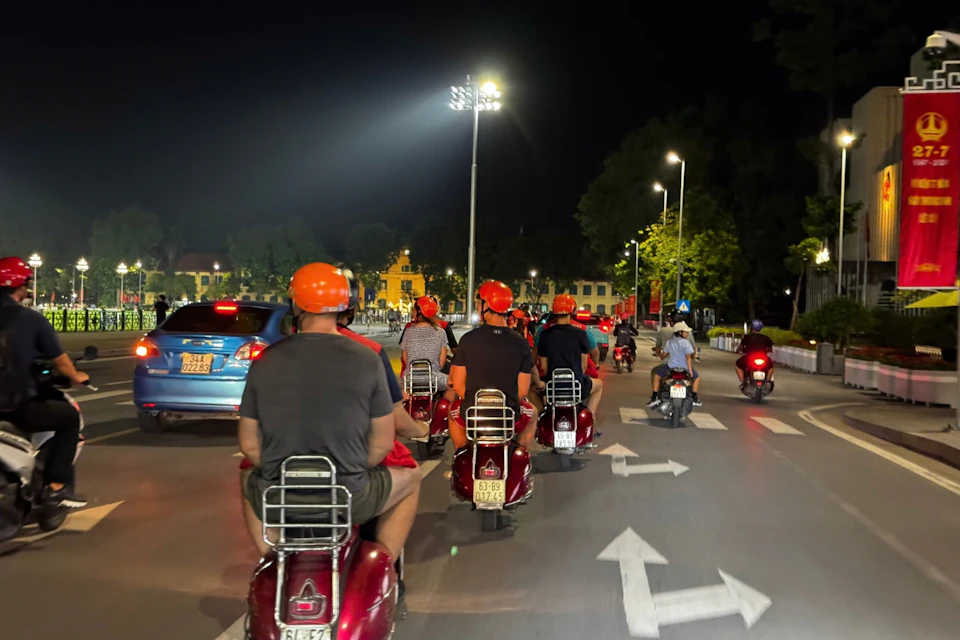 People riding red scooters with orange helmets on a lit city street at night, surrounded by other vehicles and traffic signs.