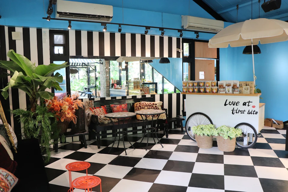 A cozy cafe interior with black and white decor, striped walls, a checkered floor, plants, and vintage-style furniture.