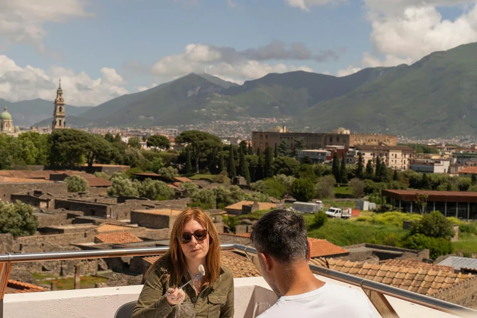 A woman and a man sit at an outdoor table overlooking an ancient ruin with mountains and a bell tower in the background.