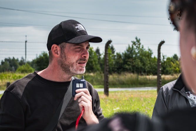 一名男子在户外手持对讲机，戴着一顶黑色帽子和衬衫，背景是绿色植物和围栏。