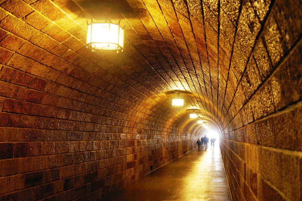 A dimly lit brick tunnel with warm lights hanging from the ceiling, ending in a bright light with silhouettes of people walking.