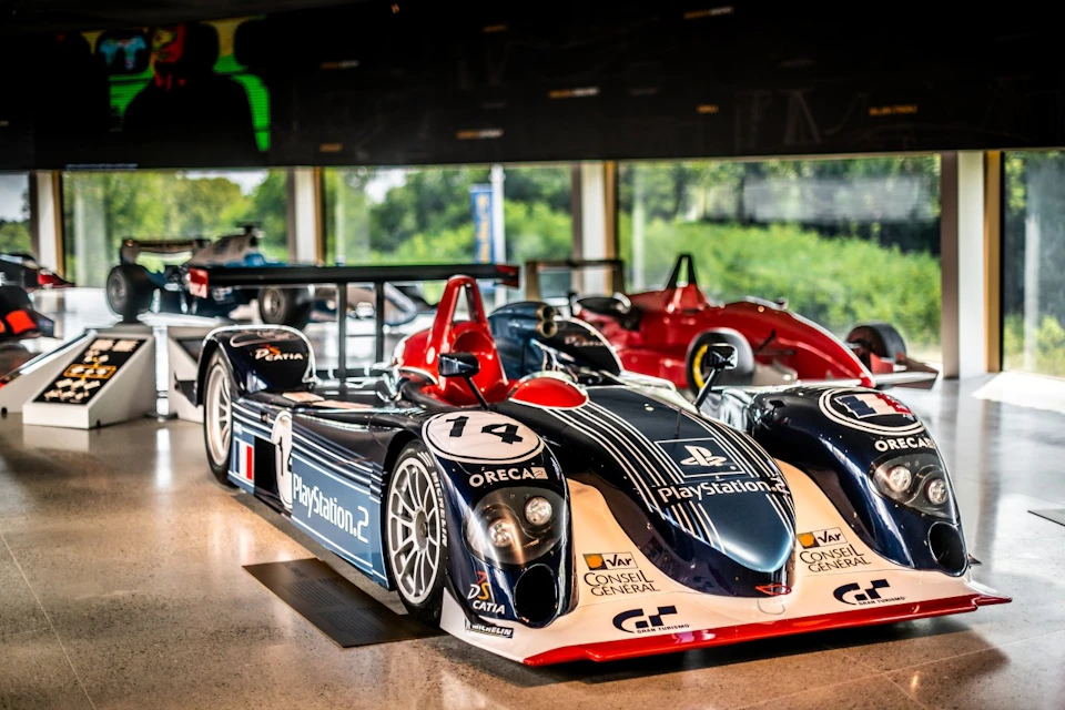 A sleek race car with PlayStation 2 branding on display indoors, surrounded by other race cars and large windows showing greenery outside.