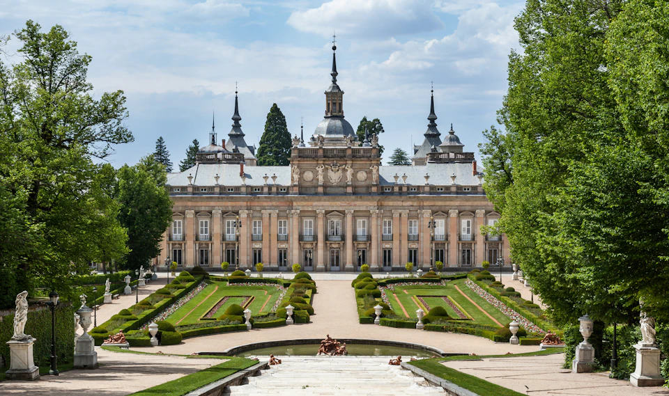 A grand palace with ornate towers and manicured gardens featuring symmetrical hedges, pathways, and a central fountain.