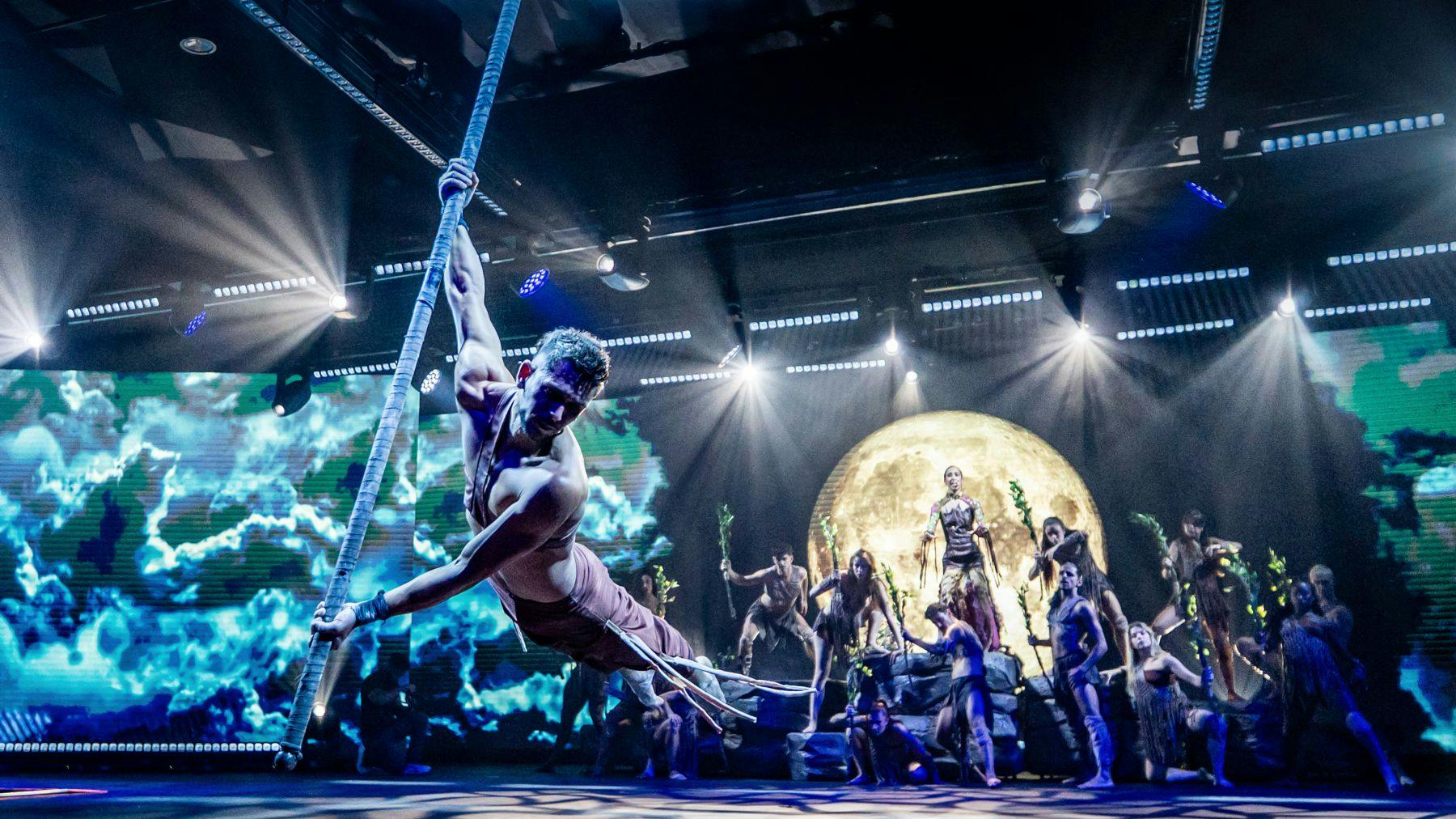 Artistas em palco num ato dramático com um pano de fundo lunar, um aerialista numa corda e outros em poses dinâmicas.