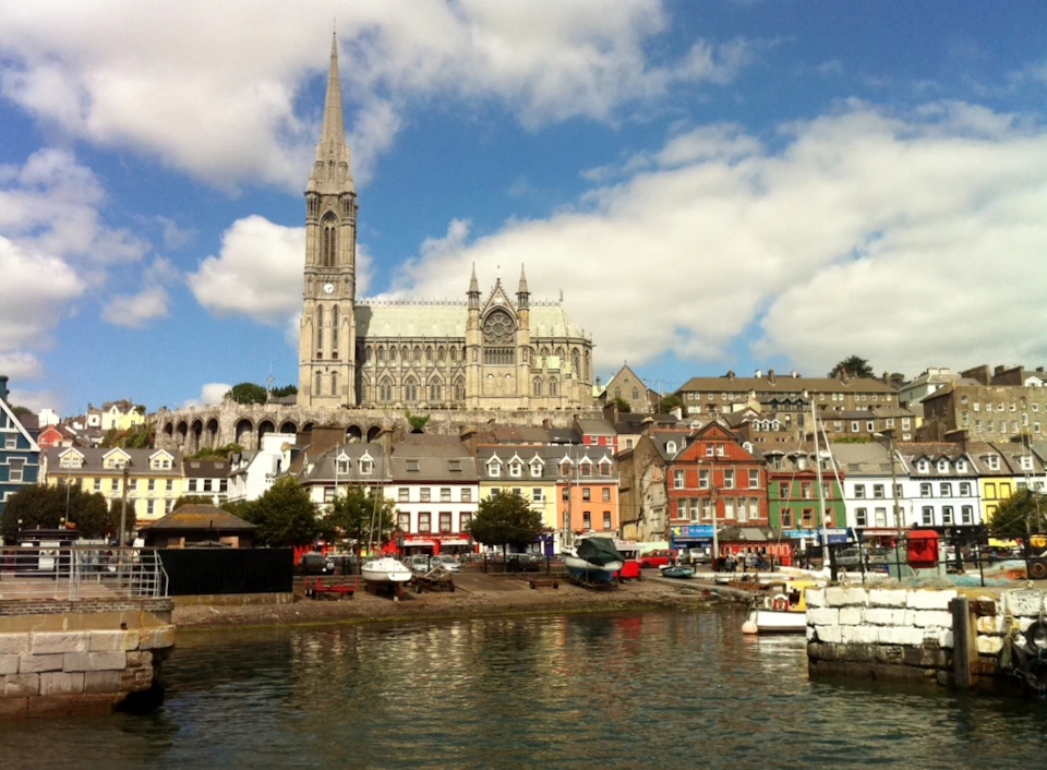 A large cathedral towers above colorful buildings and boats docked near the waterfront under a partly cloudy sky.
