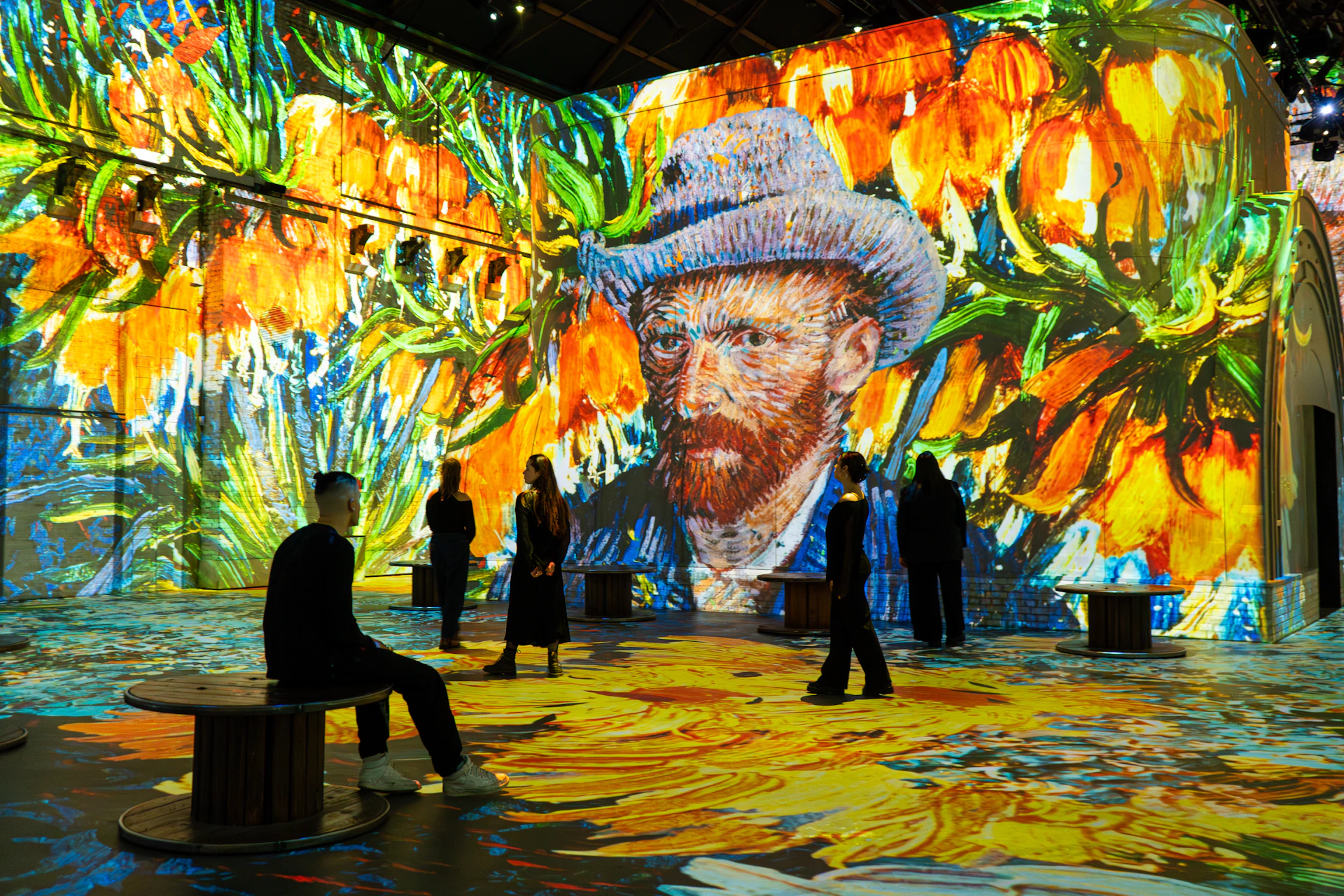 People observing a digital Van Gogh art installation with vibrant colors and large floral projections on the walls and floor.