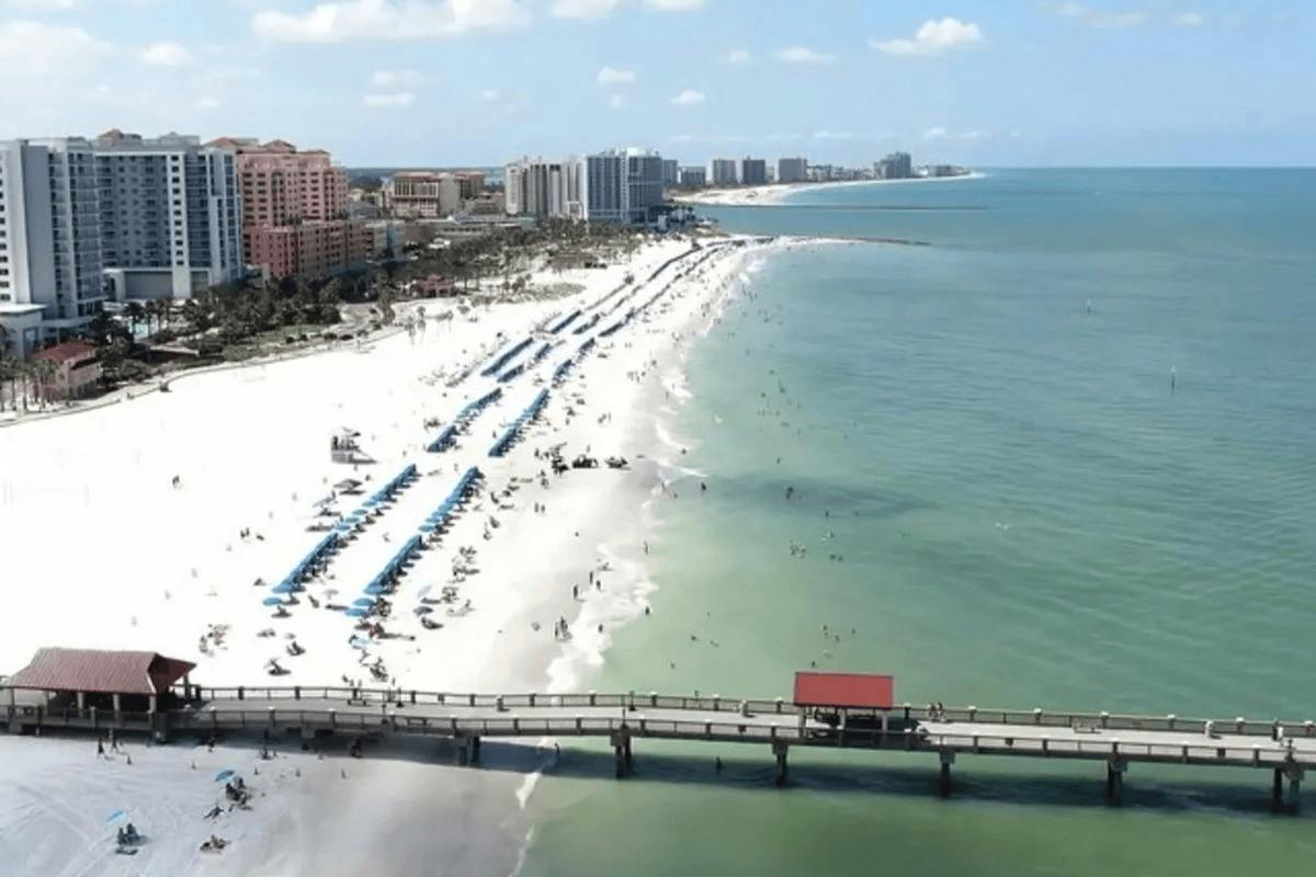 A beach with umbrellas and people alongside a pier, with buildings in the background and the ocean extending to the horizon.