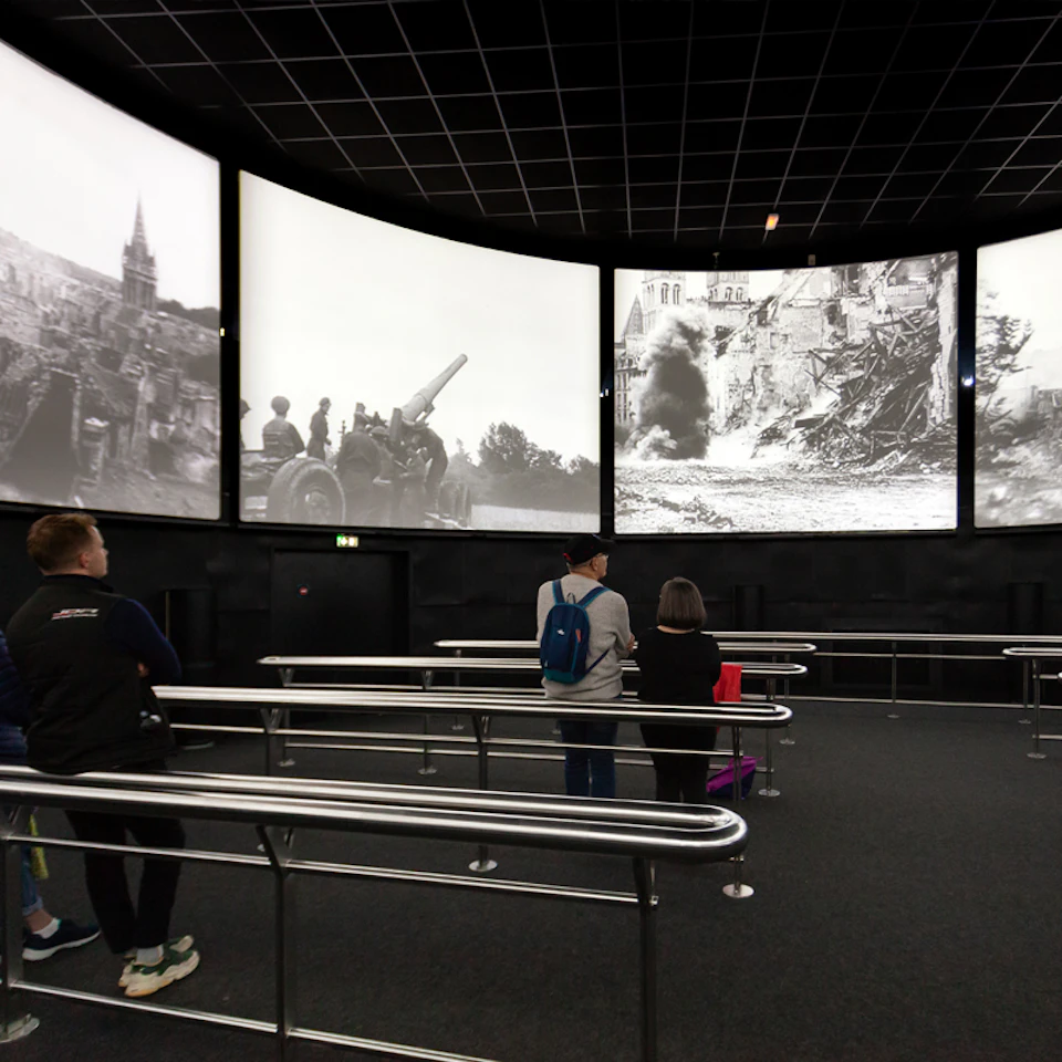 People stand in a dark room, watching World War II-era black-and-white footage displayed on large screens.