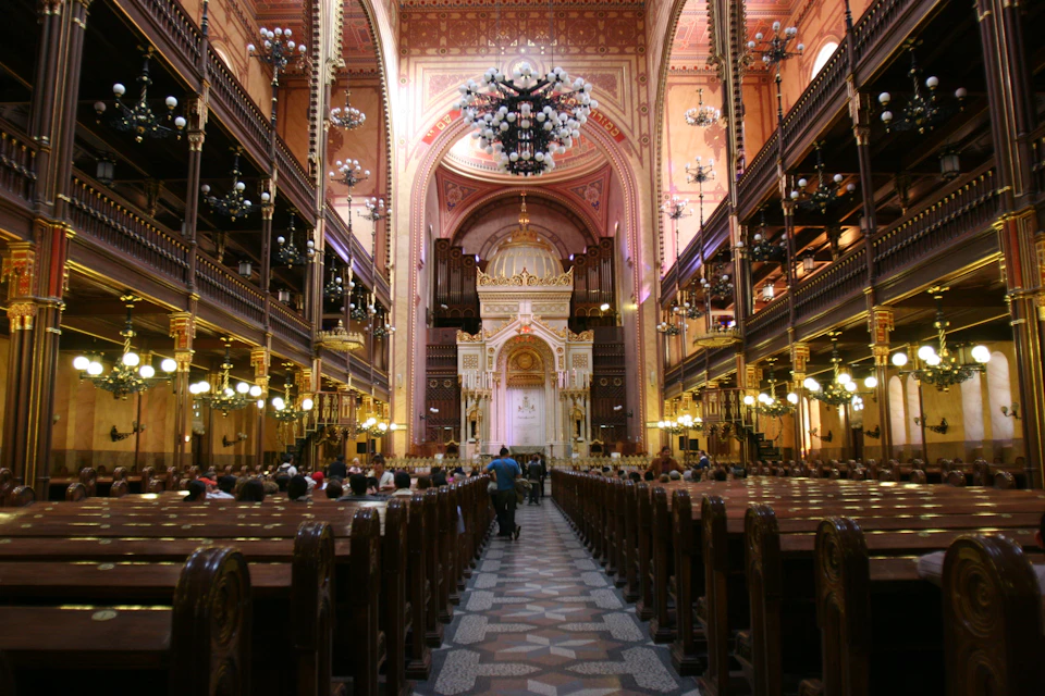 Ornate interior of a large synagogue, featuring chandeliers, intricate details, wooden pews, and people sitting and walking.