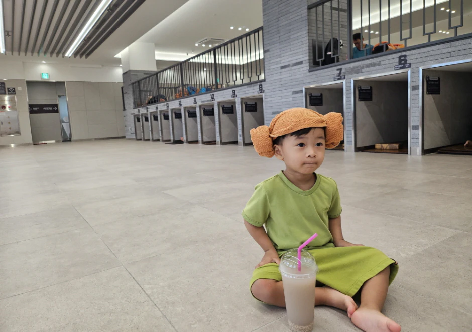 A child wearing a green outfit and an orange hat sits on the floor near a drink with a pink straw in a modern space.