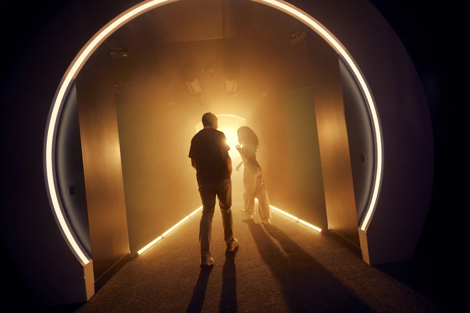 Two people walking through a dimly lit, arched corridor illuminated by warm, glowing lights.