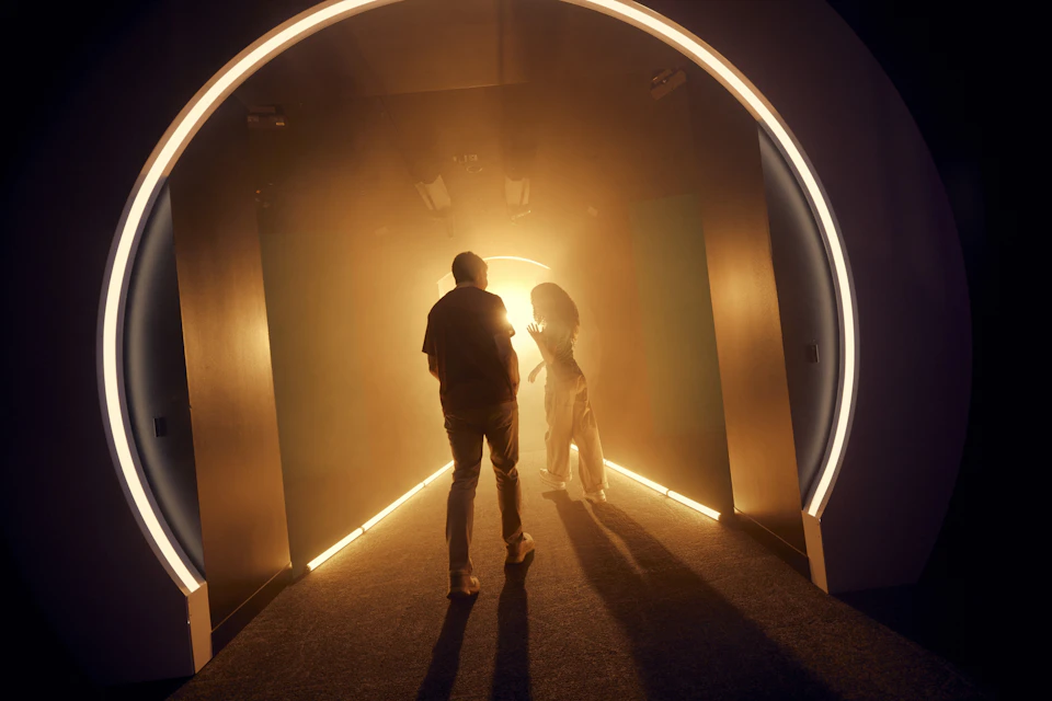 Two people walking through a dimly lit, arched corridor illuminated by warm, glowing lights.