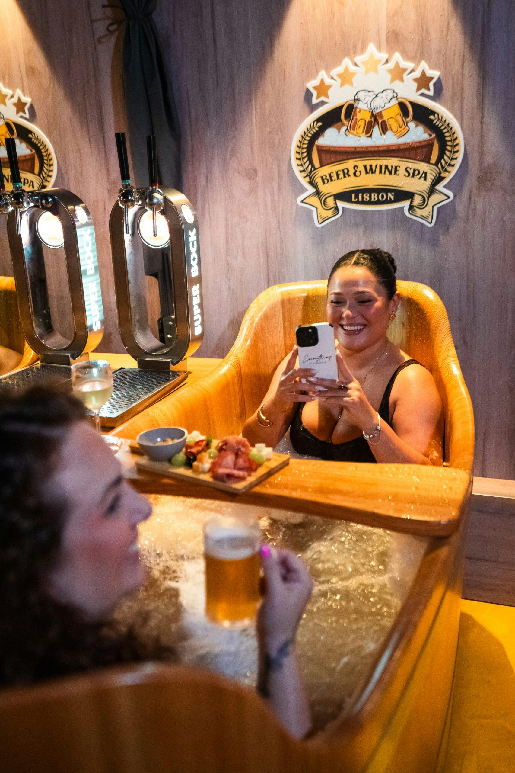 Μια γυναίκα σε μπανιέρα-υδρομασάζ χαμογελάει και κρατάει ένα τηλέφωνο. Ένα άλλο άτομο με μπύρα είναι κοντά. Πινακίδα "Beer & Wine Spa Lisbon" στο παρασκήνιο.