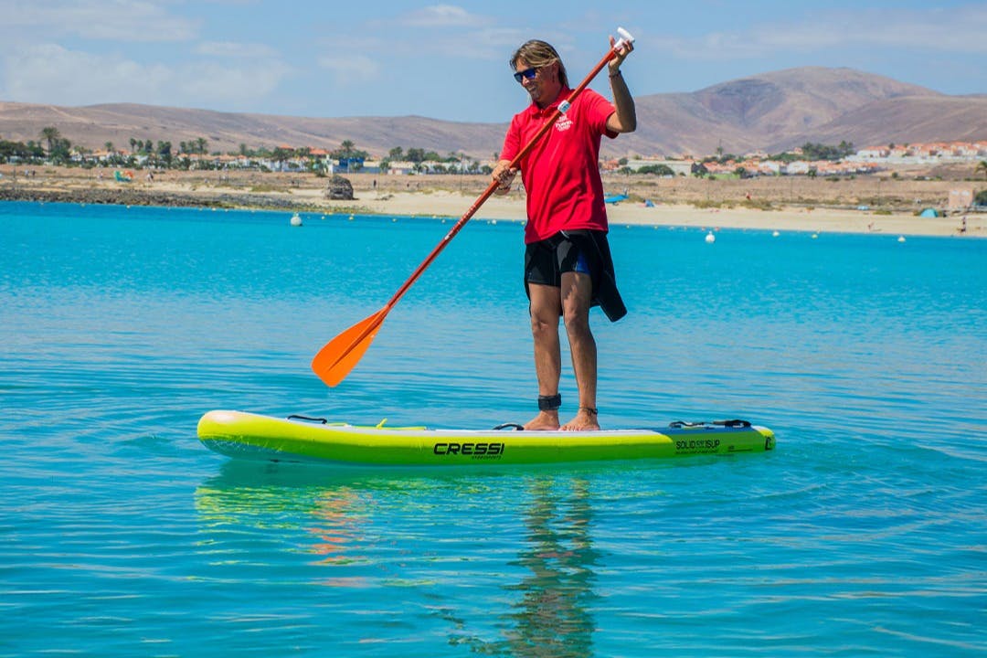 Πρόσωπο που κάνει κωπηλασία σε καταγάλανα νερά, φορώντας ένα κόκκινο πουκάμισο και μαύρο σορτσάκι, με μια παραλία και βουνά στο παρασκήνιο.