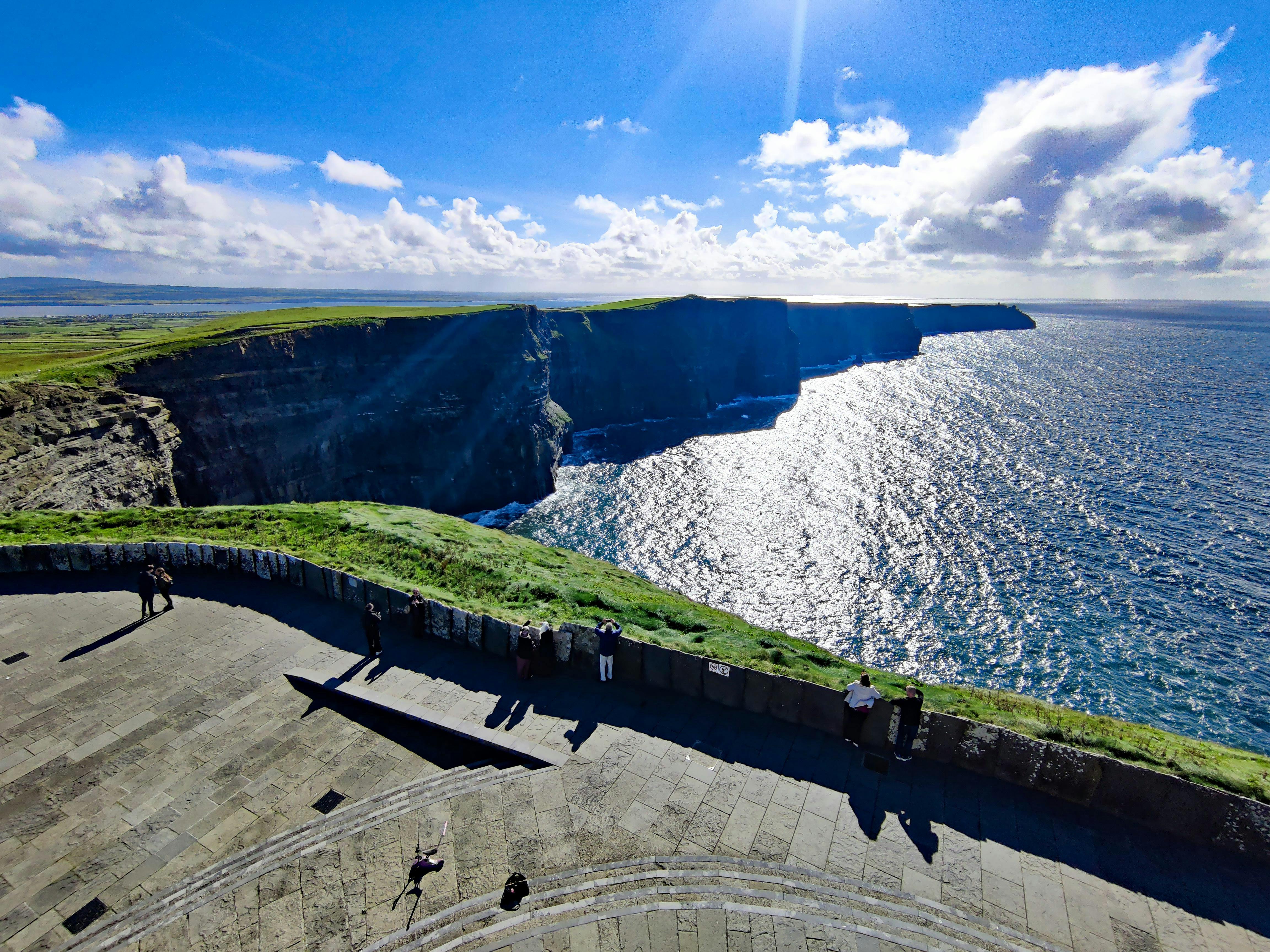 Tourists stand on a paved viewing area overlooking the Cliffs of Moher, which rise dramatically above the ocean under a bright sky.