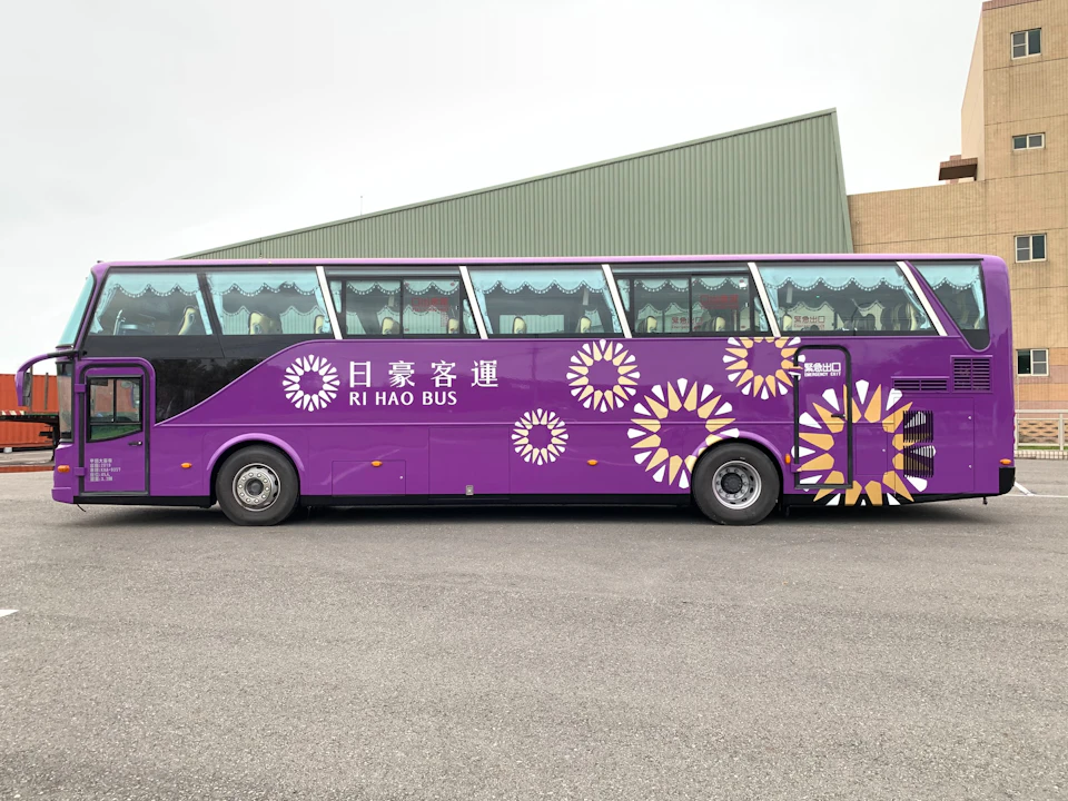 A purple bus with floral designs and text "RI HAO BUS" parked on a paved surface near a green building.