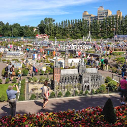 Fullsatt utomhuspark med människor som går runt detaljerade miniatyrmodeller av byggnader och landskap under en klarblå himmel.