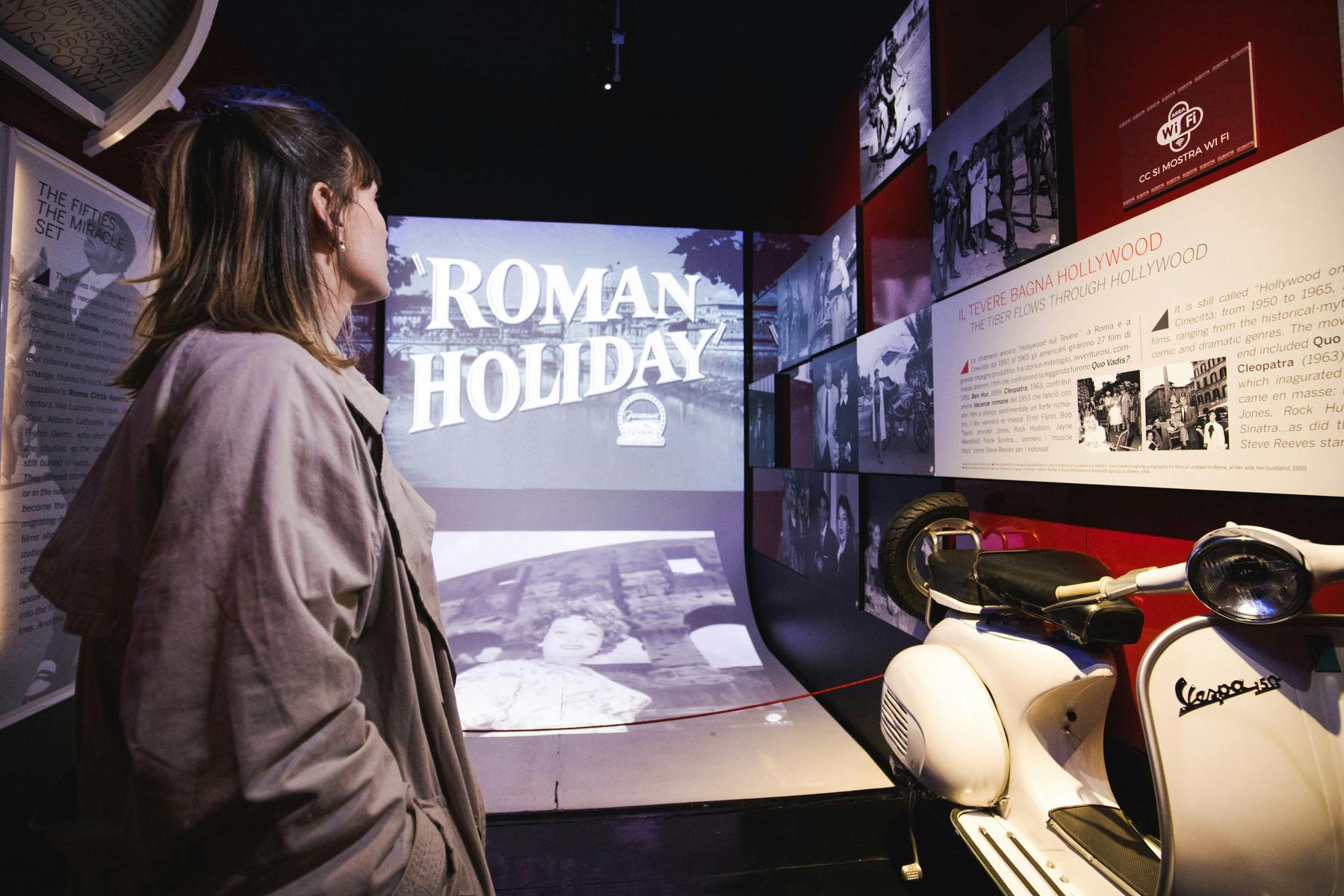 A person observes a display featuring the title 'Roman Holiday' in a museum-like setting with various images on the walls.