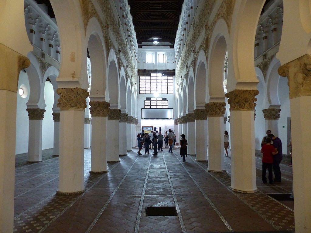 Folk, der går inde i en stor hal med hvælvede hvide søjler, indviklede udskæringer og et højt træloft.