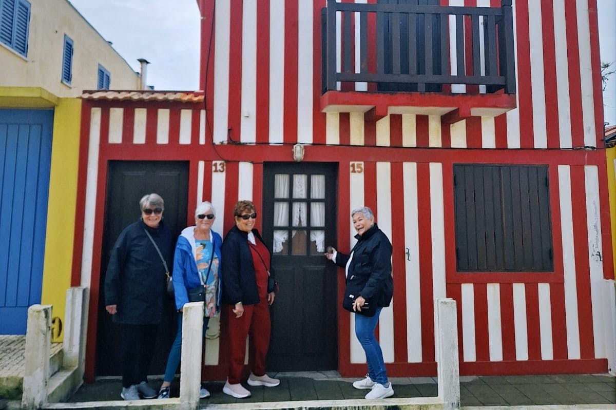 Τέσσερα άτομα στέκονται μπροστά από ένα κόκκινο και άσπρο ριγέ κτίριο, χαμογελώντας. Οι αριθμοί των σπιτιών 13 και 15 είναι ορατοί.