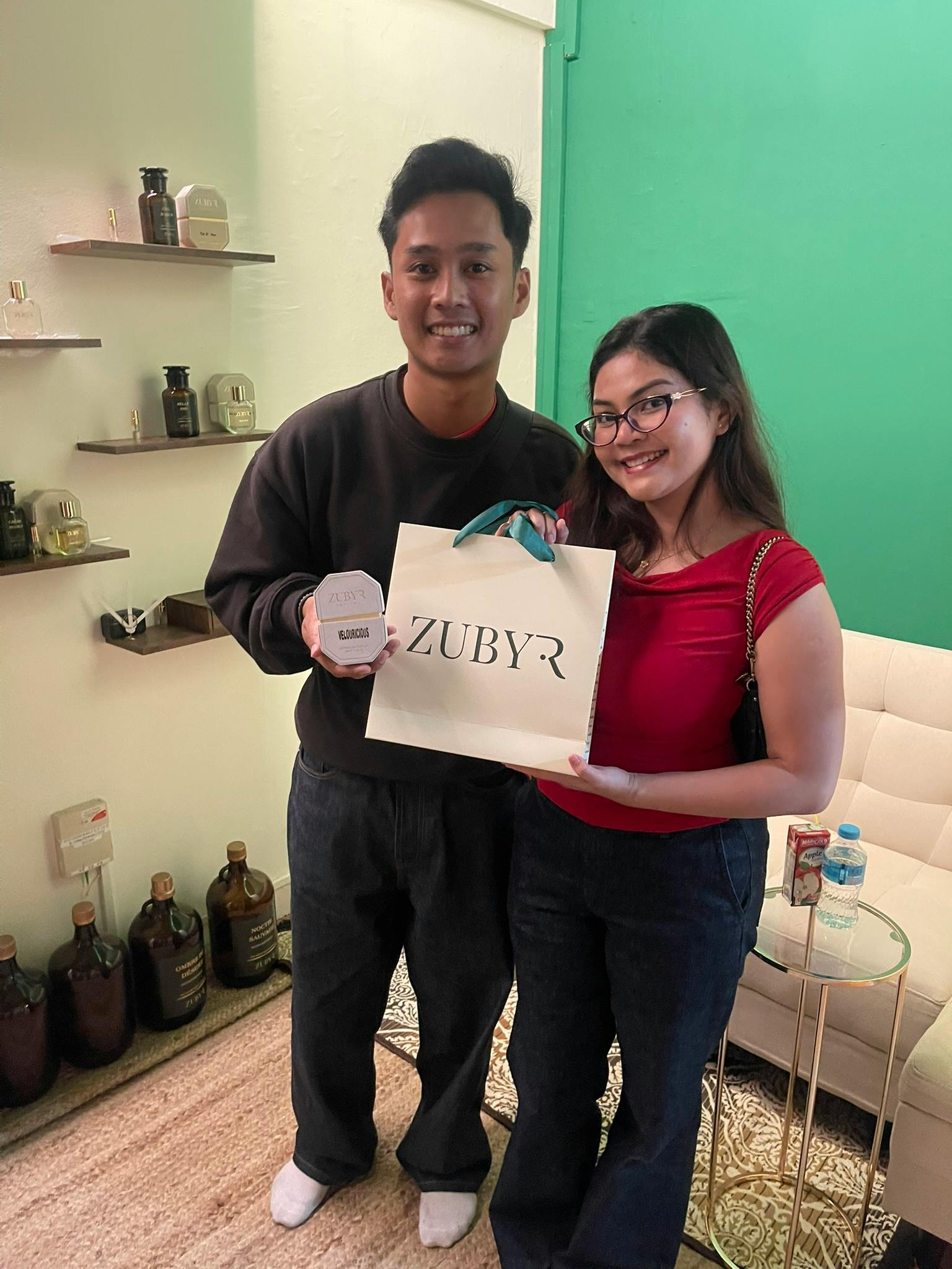 A man and a woman smiling indoors, holding a bag labeled "ZUBYR" and a small box. Shelves with bottles are in the background.