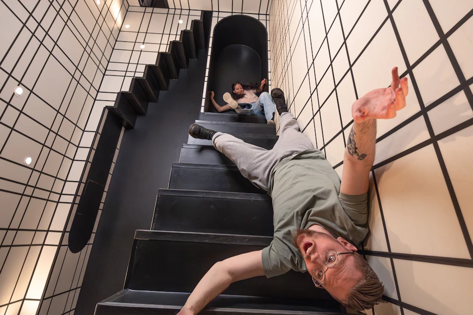 Two people appear on a staircase in a surreal perspective, with one lying down and another further up the stairs.