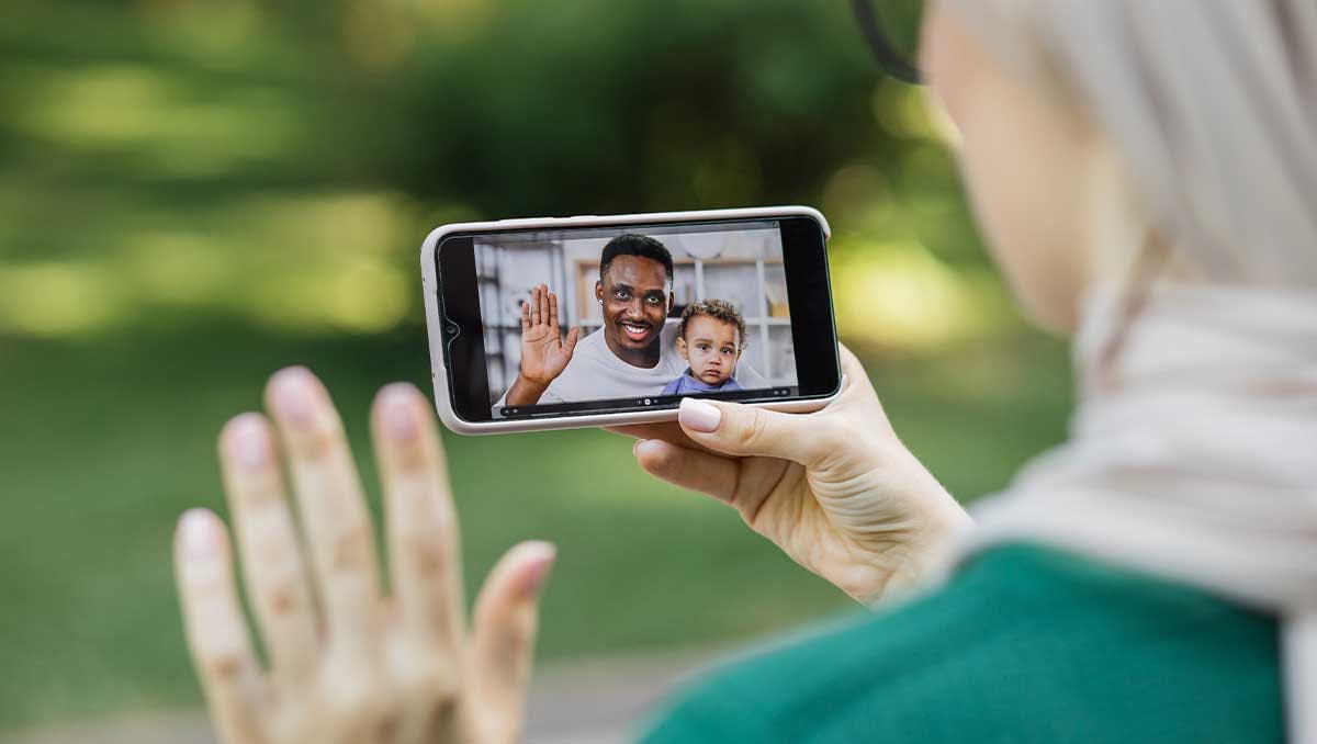 Eine Person hält ein Smartphone in der Hand, auf dem ein Videogespräch mit einem Mann und einem winkenden Kind angezeigt wird. Man sieht eine andere Hand, die zurückwinkt.