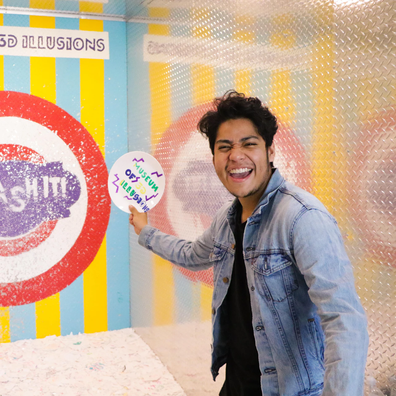 Person smiling and holding a sign in a colorful booth with "Museum of 3D Illusions" written above and other text partially visible.