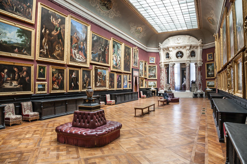 Spacious art gallery with ornate ceiling, large paintings on red walls, classic wooden flooring, and cushioned seating.